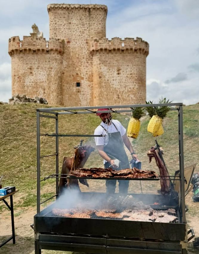Asador de El Ombú preparando carne a la cruz frente a un castillo